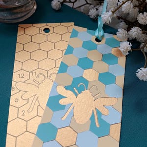 May include: Two rectangular bookmarks featuring a honeycomb pattern. One bookmark is gold with a bee outline and numbers. The other is a mix of teal, light blue, and gold hexagons with a solid gold bee silhouette and a light blue ribbon at the top.