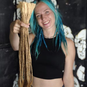 Può includere: Una persona con i capelli blu tiene in mano un pacchetto di extension per capelli dreadlocks di colore marrone chiaro. Le extension sono lunghe circa 50 cm.