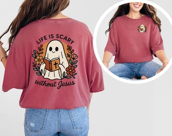 Op de afbeelding: Een stoffig roze t-shirt met een afbeelding van een vriendelijk spook dat een boek met een kruis vasthoudt. De tekst "LIFE IS SCARY without Jesus" staat boven en onder het spook. Het spook is omringd door bloemen.
