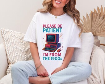 以下が含まれることがあります： レトロなコンピューターグラフィックと、ターコイズとピンクで「PLEASE BE PATIENT I'M FROM THE 1900s」というテキストがプリントされた白いTシャツ。 ヴィンテージのコンピューター、キーボード、マウスがデザインされています。