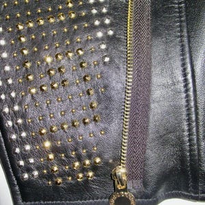 May include: Close-up of a black leather jacket with gold and silver studs. The jacket has a zipper closure with a gold zipper pull.