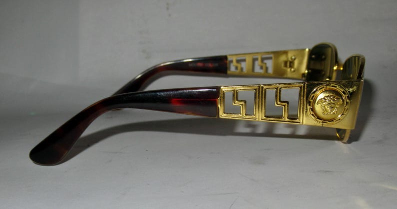 May include: Gold-tone sunglasses with a tortoiseshell frame and a Versace logo on the side. The sunglasses have a geometric design on the frame.