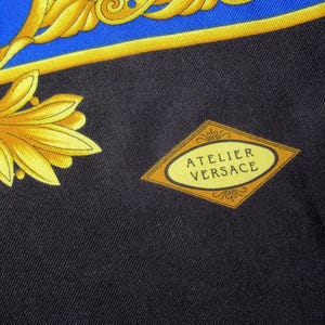 Puede incluir: Una tela negra con un patr&oacute;n dorado y azul. La tela tiene una etiqueta en forma de diamante con el texto "Atelier Versace" en letras doradas.