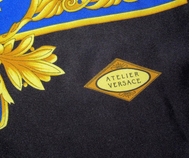Puede incluir: Una tela negra con un patr&oacute;n dorado y azul. La tela tiene una etiqueta en forma de diamante con el texto "Atelier Versace" en letras doradas.