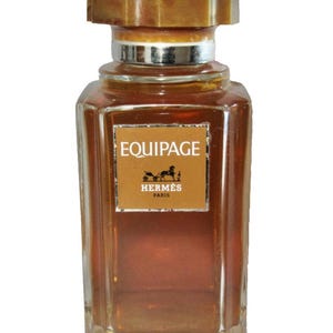 Op de afbeelding: Een rechthoekige parfumfles met een bruine vloeistof erin. De fles heeft een bruine dop en een zilveren band om de hals. Het etiket leest "EQUIPAGE" en "HERMÈS PARIS" met een paard-en-wagen-logo.