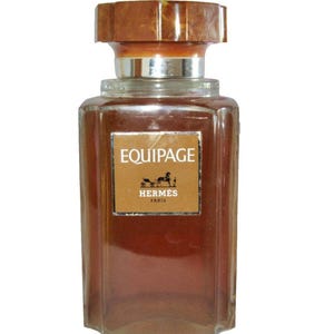 Op de afbeelding: Een rechthoekige glazen parfumfles met een bruine dop en een zilveren hals. De fles heeft een bruin etiket met het woord "EQUIPAGE" en het Hermes-logo. De parfum in de fles is lichtbruin van kleur.