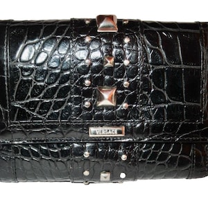 May include: Black wallet with a crocodile skin texture, adorned with silver-toned studs and square accents. The wallet features a rectangular silver plaque with the brand name "VERSACE".