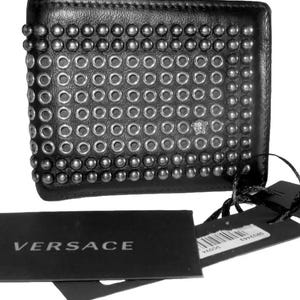 Może przedstawiać: Czarny skórzany portfel z srebrnymi ćwiekami i oczkami. Portfel ma napis "VERSACE" nadrukowany na czarnej metce.