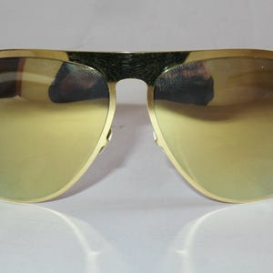 Puede incluir: Gafas de sol doradas de aviador con lentes espejadas. Las gafas tienen una forma clásica de lágrima y una montura de metal dorado. Las lentes reflejan un tono amarillo-dorado. El puente de las gafas es oscuro.