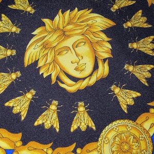 Puede incluir: Una tela azul oscuro con un dise&ntilde;o dorado y amarillo. El dise&ntilde;o presenta la cara de una mujer con una corona de hojas y serpientes, rodeada de abejas.