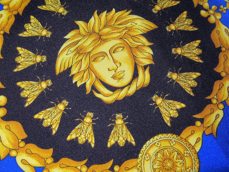 Puede incluir: Una tela azul oscuro con un dise&ntilde;o dorado y amarillo. El dise&ntilde;o presenta la cara de una mujer con una corona de hojas y serpientes, rodeada de abejas.