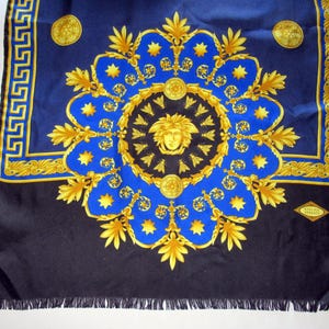 Puede incluir: Un pa&ntilde;uelo de seda negro con un patr&oacute;n azul y dorado. El patr&oacute;n presenta un medall&oacute;n central con una cara estilizada y un motivo repetitivo de hojas y estrellas. El pa&ntilde;uelo tiene un borde de flecos y una peque&ntilde;a etiqueta que dice "Gianni Versace".