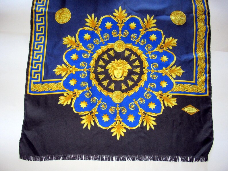 Puede incluir: Un pa&ntilde;uelo de seda negro con un patr&oacute;n azul y dorado. El patr&oacute;n presenta un medall&oacute;n central con una cara estilizada y un motivo repetitivo de hojas y estrellas. El pa&ntilde;uelo tiene un borde de flecos y una peque&ntilde;a etiqueta que dice "Gianni Versace".