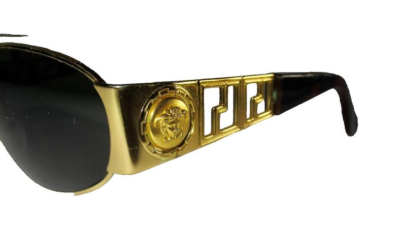 May include: Gold-tone sunglasses with a black lens and a Versace logo on the side. The sunglasses have a brown plastic arm.