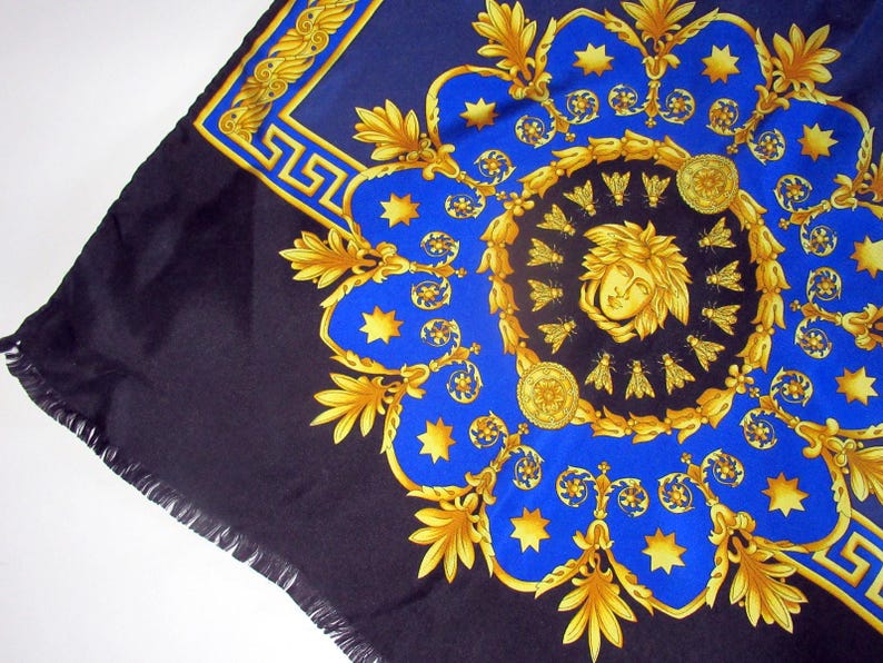 Puede incluir: Un pa&ntilde;uelo de seda negro y azul con un patr&oacute;n dorado y azul. El patr&oacute;n presenta un medall&oacute;n central con una cara estilizada y abejas, rodeado de un dise&ntilde;o repetitivo de estrellas, hojas y volutas.