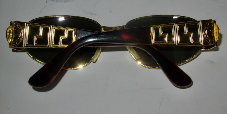 May include: Gold-tone sunglasses with black frames and dark lenses. The arms of the sunglasses have a gold-tone pattern.