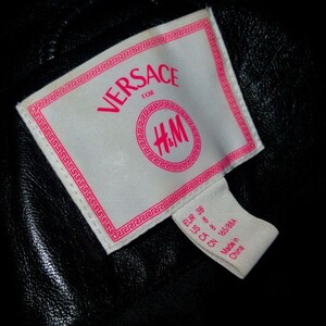 May include: A white tag with pink text that reads "VERSACE for H&M". The tag has a Greek key border. A smaller white tag with pink text indicates the size: "EUR 38 US 8 CA 10 665884 Made in China."