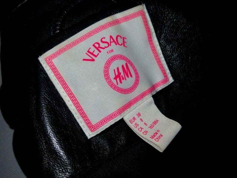 May include: A white tag with pink text that reads "VERSACE for H&M". The tag has a Greek key border. A smaller white tag with pink text indicates the size: "EUR 38 US 8 CA 10 665884 Made in China."