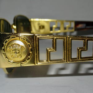 May include: Gold-tone sunglasses with a Medusa head design on the side and a Greek key pattern on the frame.