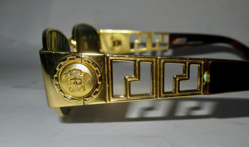 May include: Gold-tone sunglasses with a Medusa head design on the side and a Greek key pattern on the frame.