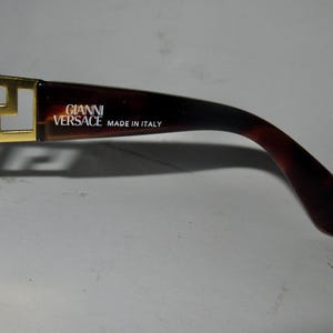 May include: Gold-tone sunglasses with a brown tortoiseshell frame. The frame has the text "GIANNI VERSACE MADE IN ITALY" printed on it.