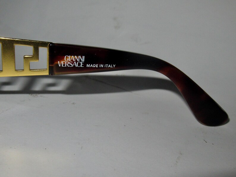 May include: Gold-tone sunglasses with a brown tortoiseshell frame. The frame has the text "GIANNI VERSACE MADE IN ITALY" printed on it.