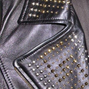 May include: Black leather jacket with a studded collar and a zip closure. The collar and the front flap are decorated with silver and gold studs.
