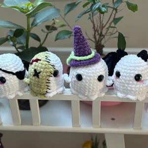 May include: Four crocheted ghost ornaments with different Halloween themes. One ghost wears a purple hat, one has a stitched-up face, one has a black eye patch, and one has black bat wings.