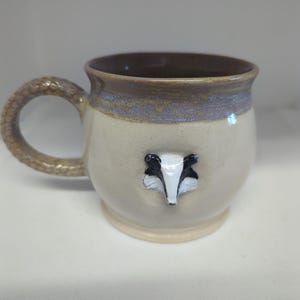 May include: A ceramic mug with a cream-colored body and a brown interior. The rim features a band of mottled purple and brown glaze. A sculpted badger face is attached to the front, and the handle has a textured, scale-like pattern.