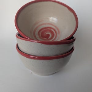 Puede incluir: Una pila de tres cuencos de cerámica. Los cuencos son de color blanquecino con una textura moteada y un diseño de espiral rojo en el centro. Los bordes también son rojos.