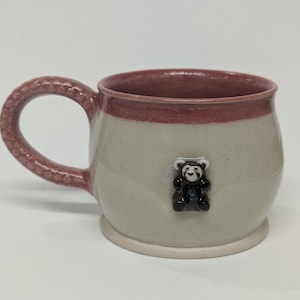 Könnte beinhalten: Handgefertigte Keramiktasse mit hellbeigem Korpus und dunkelrosa Rand und Henkel. Eine kleine, dunkelbraune Teddybär-Figur ist an der Vorderseite der Tasse befestigt. Der Griff hat ein strukturiertes, schuppenartiges Design.