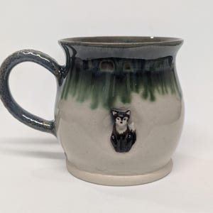 Peut inclure: Une tasse en céramique avec une anse et un bord bleu foncé, présentant un corps beige clair avec un glaçage vert foncé et bleu. Une petite figurine de renard détaillée est fixée à l'avant de la tasse. La tasse est conçue pour les boissons chaudes.
