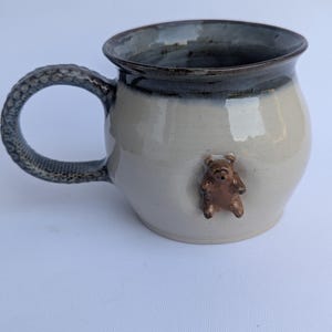 May include: A ceramic mug with a rounded body, featuring a glossy cream-coloured glaze on the main surface and a dark charcoal rim. A small, sculpted brown bear is attached to the side, and the handle has a textured, dark grey scale-like pattern.