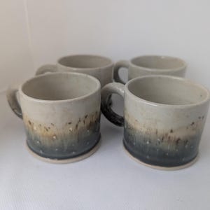 Könnte beinhalten: Vier Keramikbecher mit einem gesprenkelten Farbverlauf. Die Becher gehen von hellem Beige oben zu einem dunkelblauen Grau an der Basis über. Jeder Becher hat einen abgerundeten Henkel und eine leicht strukturierte Oberfläche.