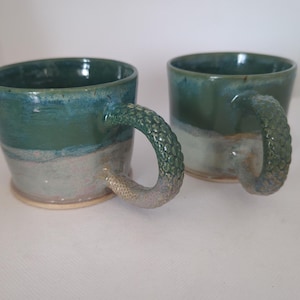 May include: Two ceramic mugs featuring a deep green and blue glazed upper section that transitions into a lighter, muted grey-blue base. Each mug includes a distinct handle textured with a raised, repeating scale pattern resembling reptile skin.