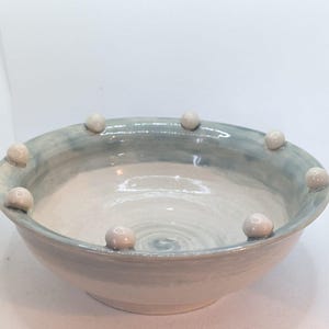 Könnte beinhalten: Eine handgefertigte Keramikschale mit hellblauer und weißer Glasur. Der Rand der Schale ist mit kleinen, runden, cremefarbenen Keramikakzenten verziert. Die Schale hat ein schlichtes, elegantes Design, geeignet zum Servieren oder Ausstellen.