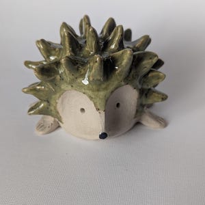 Könnte beinhalten: Keramik-Igel-Figur mit hellbeigem Gesicht und dunkelgrünem, stacheligem Rücken. Der Igel hat kleine, runde Augen und eine winzige schwarze Nase. Die Figur ist ein dekoratives Objekt.