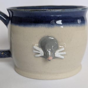 Può includere: Una tazza in ceramica con corpo color crema e bordo smaltato blu scuro. Una figura tridimensionale di una talpa grigia con zampe bianche e naso rosa è applicata al centro della tazza. Il manico è blu scuro con piccole macchie bianche.