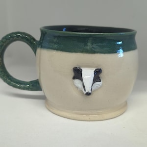May include: A ceramic mug with a cream-coloured body and a dark green glazed rim. The mug features a sculpted, three-dimensional badger face in black and white on the front and a textured, braided-style handle in the same dark green glaze.