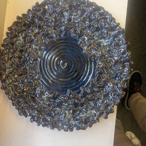 May include: A circular ceramic plate featuring a deep blue centre with a prominent spiral pattern. The outer rim is characterised by a highly textured, ruffled, and crinkled surface in shades of dark blue and black with subtle metallic highlights.