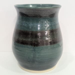May include: A ceramic vase with a glossy finish, featuring horizontal bands of deep teal, dark charcoal, and lighter blue-green. The vessel has a wide, flared rim, a rounded body that tapers towards the base, and a visible unglazed light beige bottom.