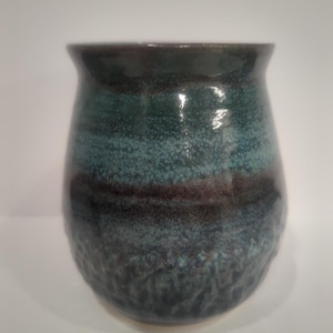 May include: A ceramic mug with a rounded, bulbous shape and a glossy finish. The surface features horizontal bands of deep teal, dark navy blue, and charcoal grey, with a speckled, mottled texture throughout the glaze that varies in colour intensity.