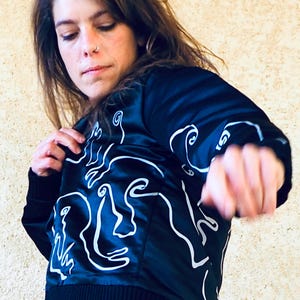 May include: A black satin bomber jacket with a white abstract design of faces. The jacket has a ribbed collar, cuffs, and hem.