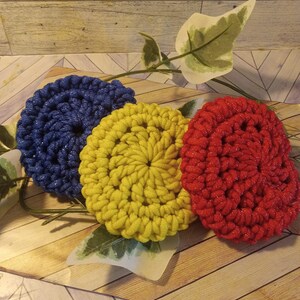 May include: Three crocheted circles in blue, yellow, and red. The circles are on a wooden surface with a green leaf.