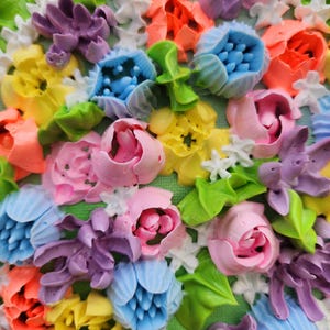 May include: A textured wall art piece featuring a dense arrangement of three-dimensional flowers in vibrant shades of pink, yellow, blue, orange, and purple. The floral composition includes sculpted petals and green leaves on a green canvas base.
