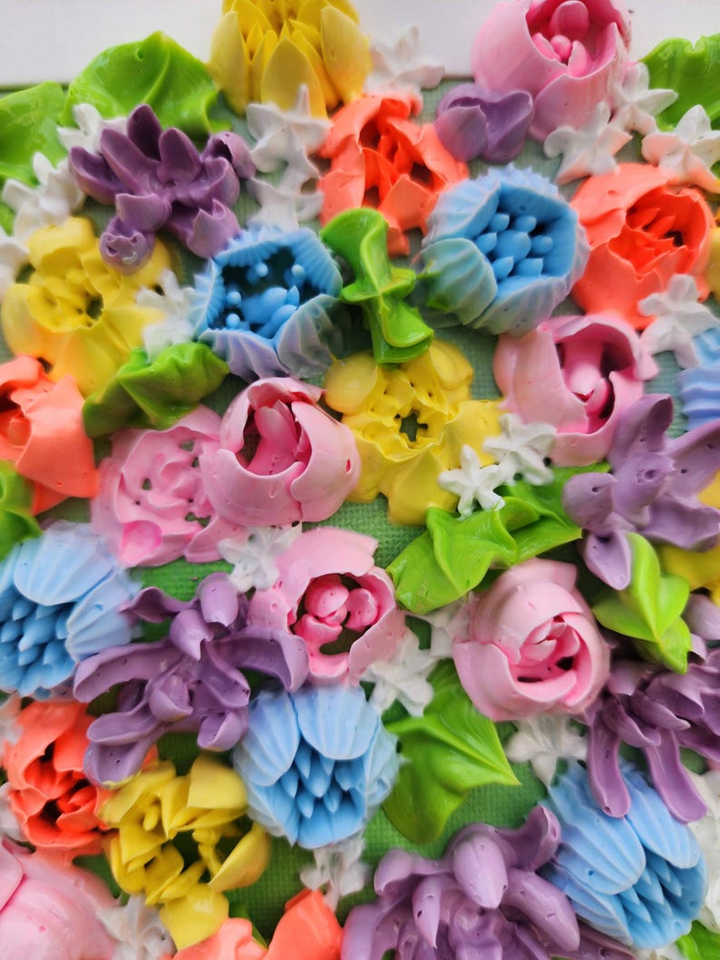 May include: A textured wall art piece featuring a dense arrangement of three-dimensional flowers in vibrant shades of pink, yellow, blue, orange, and purple. The floral composition includes sculpted petals and green leaves on a green canvas base.