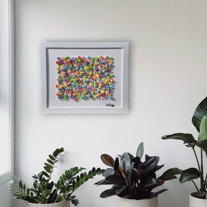 May include: A rectangular white-framed wall art piece featuring a dense, textured arrangement of small, multi-colored sculpted flowers in shades of pink, blue, yellow, purple, and green, with a signature visible in the bottom right corner.