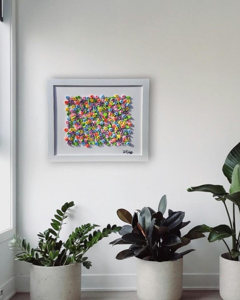 May include: A rectangular white-framed wall art piece featuring a dense, textured arrangement of small, multi-colored sculpted flowers in shades of pink, blue, yellow, purple, and green, with a signature visible in the bottom right corner.