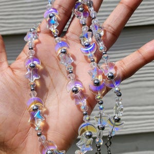 May include: A clear beaded face mask chain with iridescent star and moon charms. The chain features small, clear beads and silver-colored metal accents. The charms have a pearlescent finish, reflecting colors of purple, blue, and yellow.