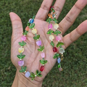 May include: A colorful beaded necklace featuring tulip-shaped beads in various colors, including pink, yellow, and blue. The necklace has green leaf accents and small silver beads. The background is a blurred green.